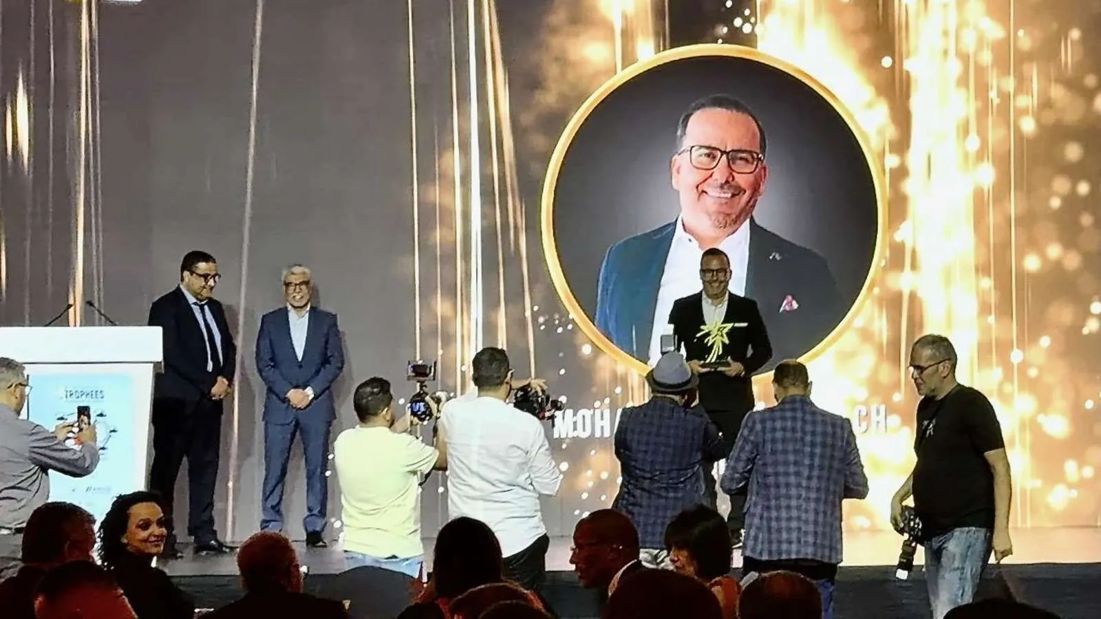 Stage of the award ceremony in Marrakech: Mohammadi Akhabach with the trophy in front of a large screen showing his portrait, surrounded by photographers.