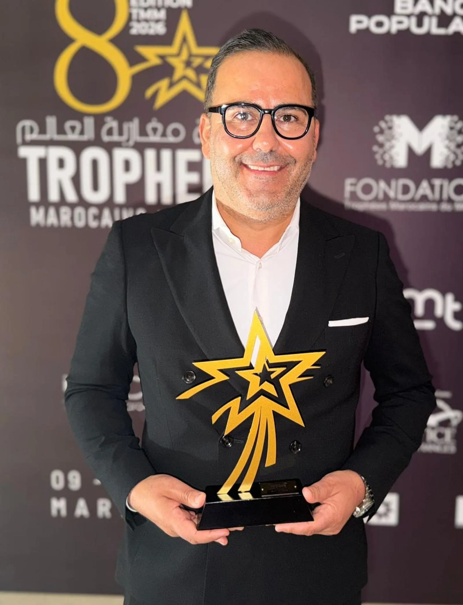 Mohammadi Akhabach in a dark suit, holding the golden star-shaped Trophées Marocains du Monde trophy in front of the event’s partner wall.
